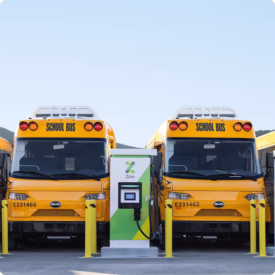 Two yellow school buses parked at an EV charging station in a bus yard, labeled "SCHOOL BUS."