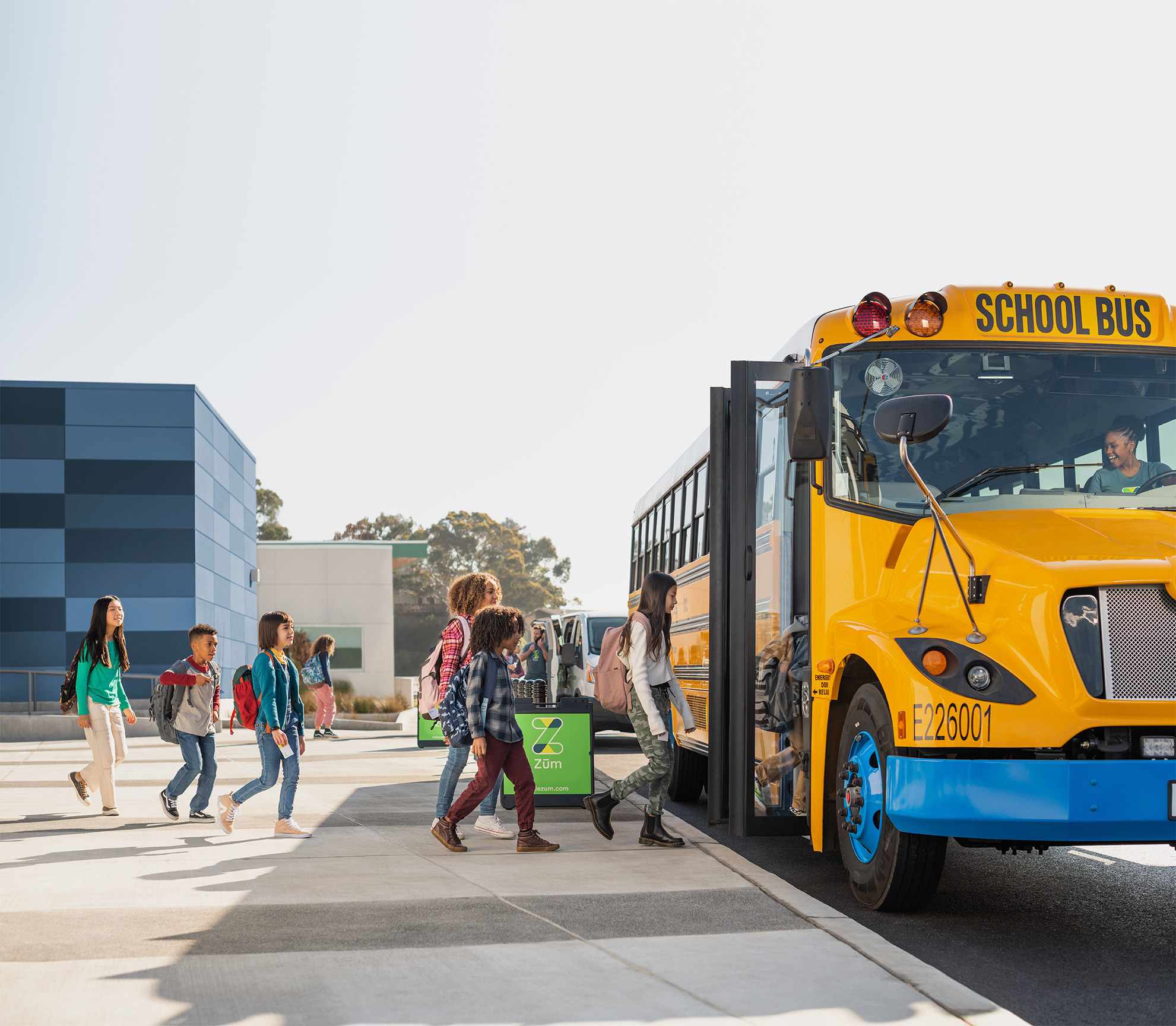 Students board a yellow school bus at a transportation hub with two buildings in view.