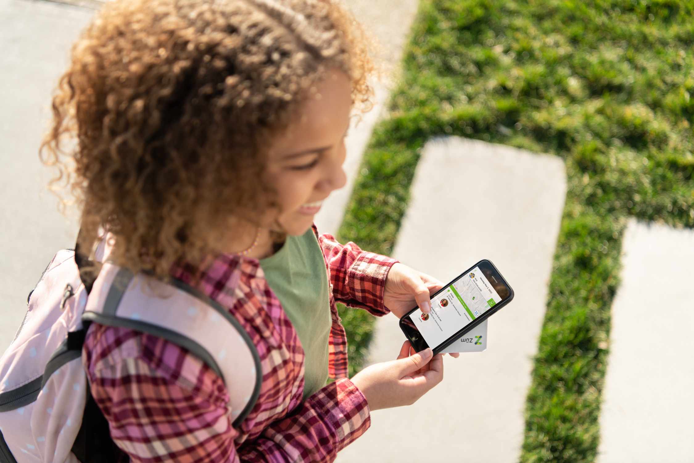 A student holds a smartphone displaying a transportation app while walking on a pathway.