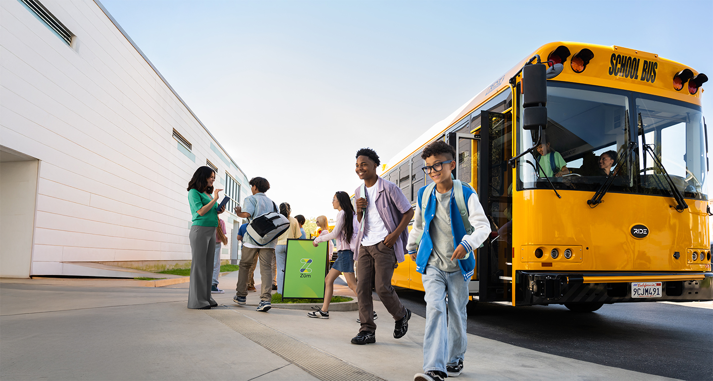 Students exiting a yellow school bus, with a school building and parents nearby.
