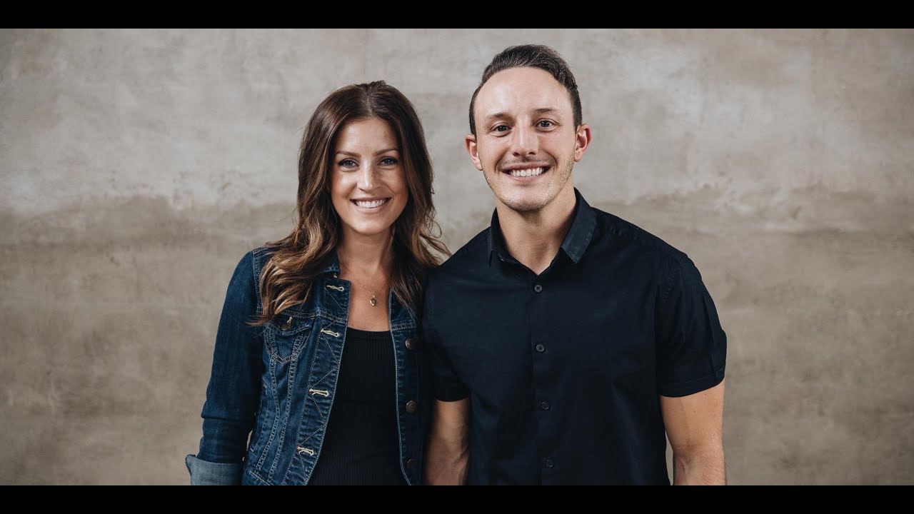 Smiling man in black shirt and woman in denim jacket stand together against gray wall.