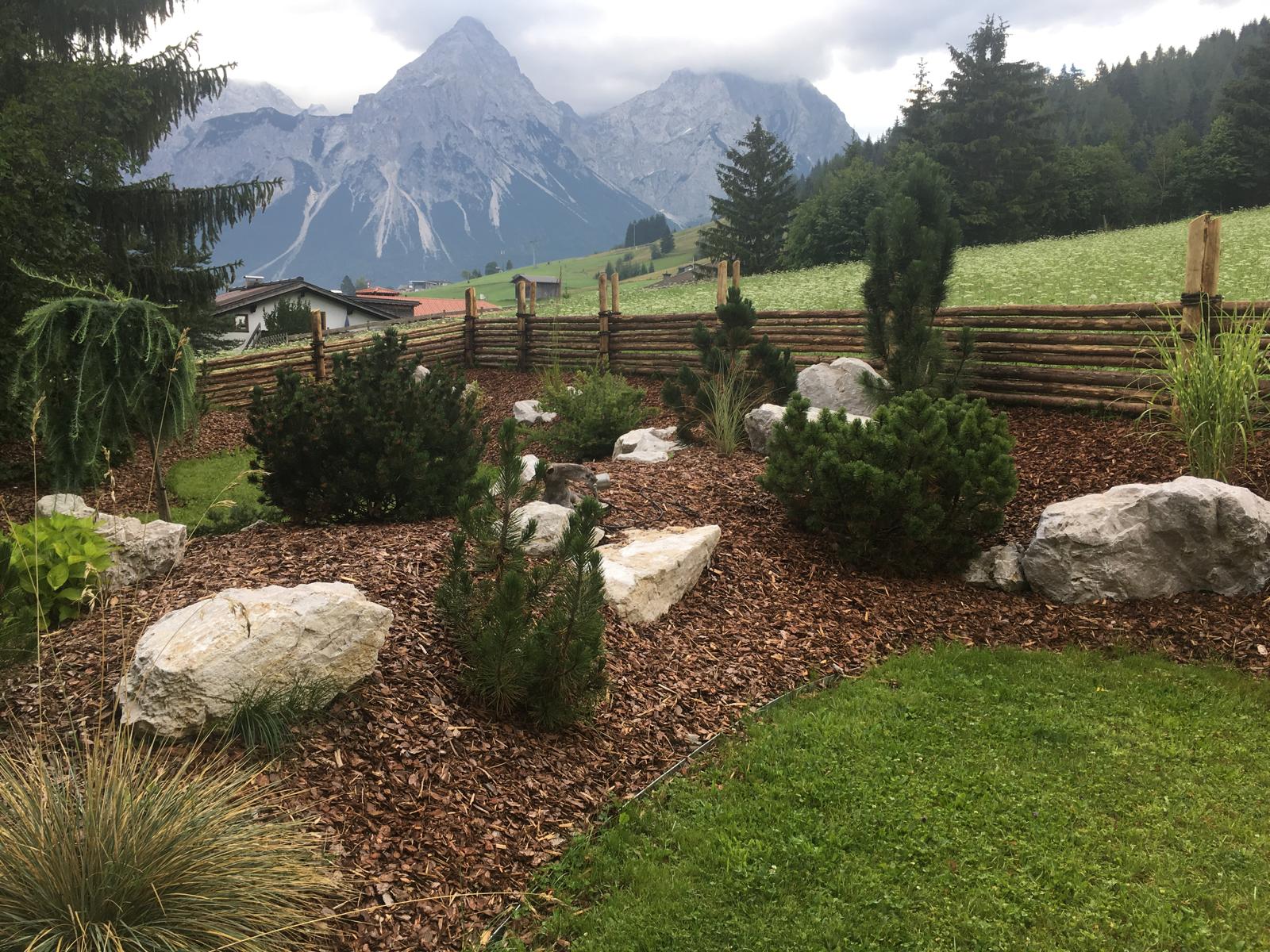 Garten mit Kieselboden, kleinen Nadelbäumen, Felsen und Holzzaun vor bewölkten Bergen.