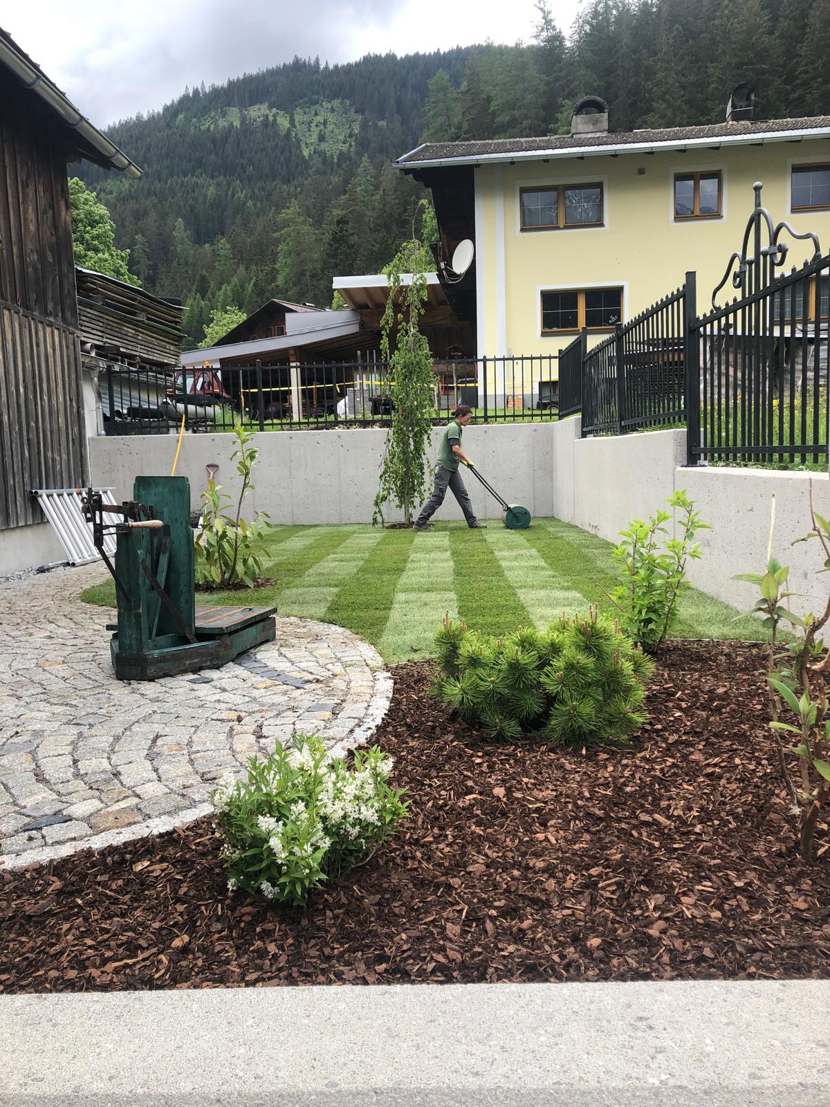 Person mäht gepflegten Rasen in einem Garten neben einem Haus mit schwarzen Zäunen und Bergen im Hintergrund.