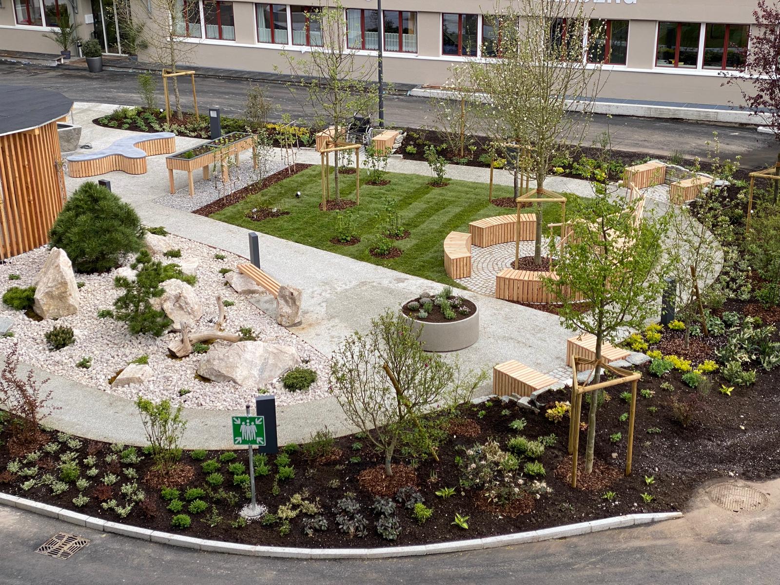 Modern gestalteter Garten mit Wegen, Sitzbänken, Bäumen und steiniger Landschaft neben einem Gebäude.