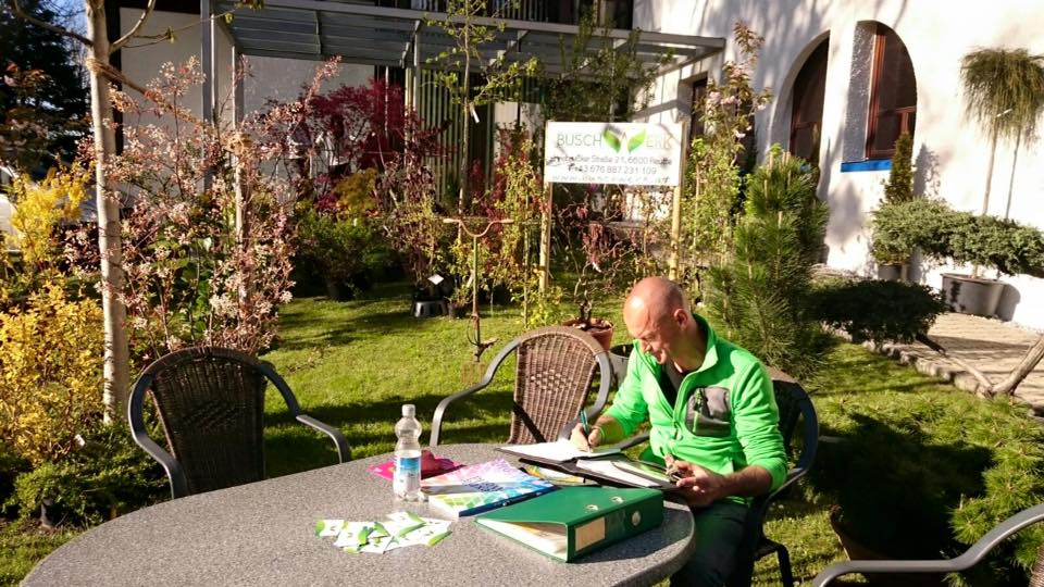 Mann schreibt Notizen an einem Gartentisch mit Ordner und Flyern vor ihm in einer hellen Gartenanlage.