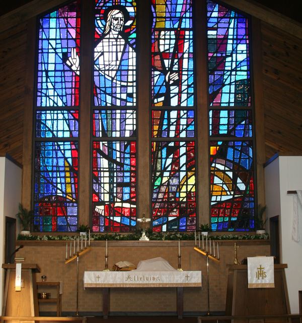 The altar and stained glass at Faith Lutheran Church in Derby, Kansas. The altar is dressed in white paraments, with a set of candles at either side. The stained glass in the background has Jesus surrounded by vibrant blues and reds. 