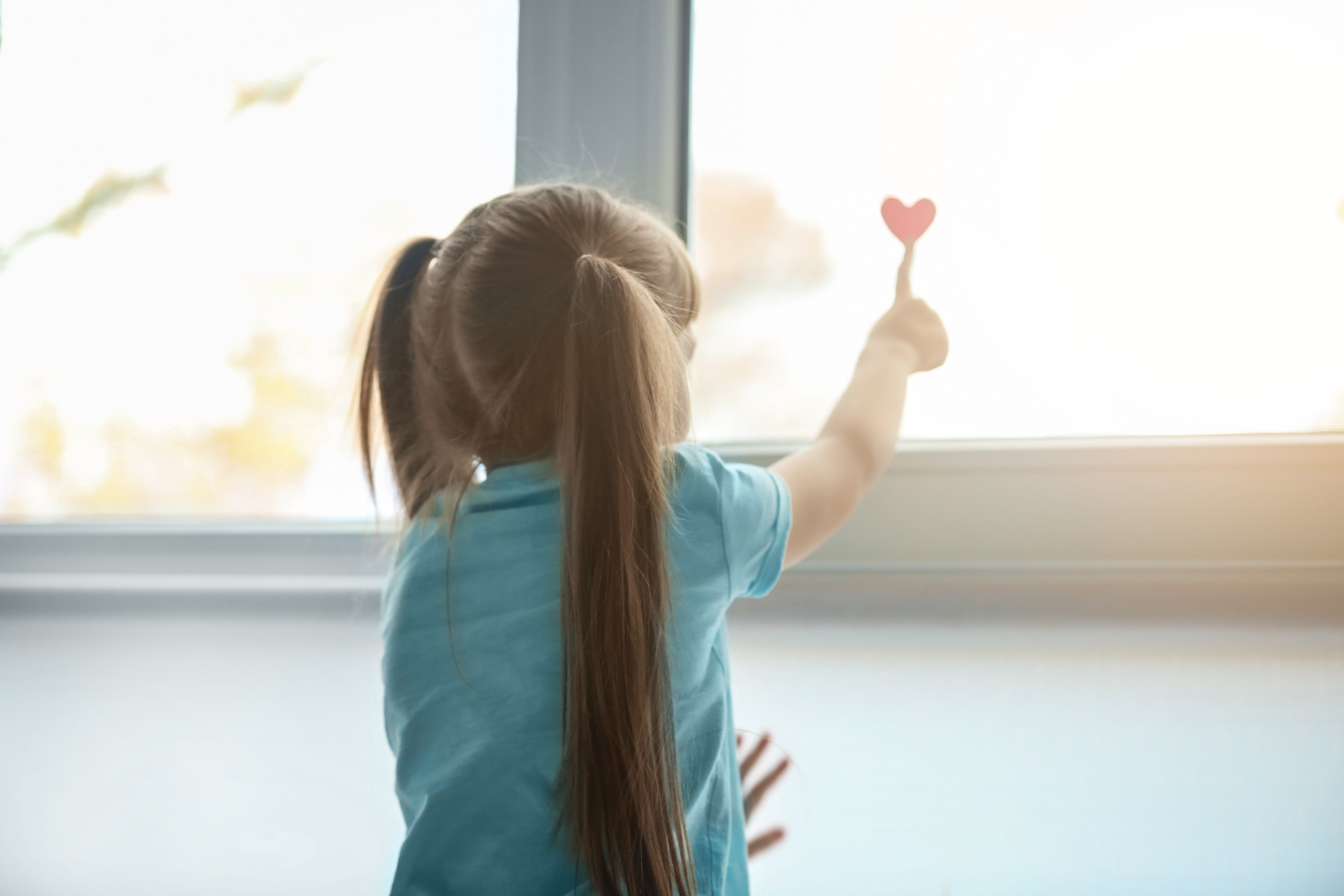 Child with long hair holding small red heart while looking out window