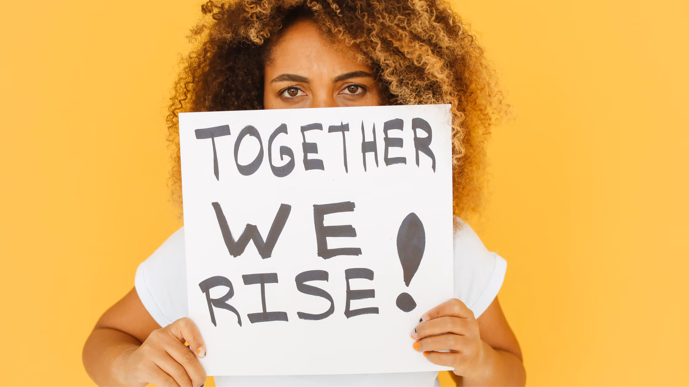 Person with curly hair holding sign reading "Together We Rise" against yellow background