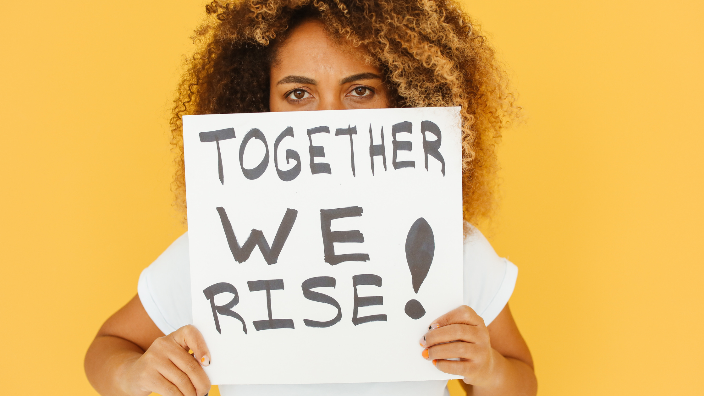 Person with curly hair holding sign reading "Together We Rise" against yellow background