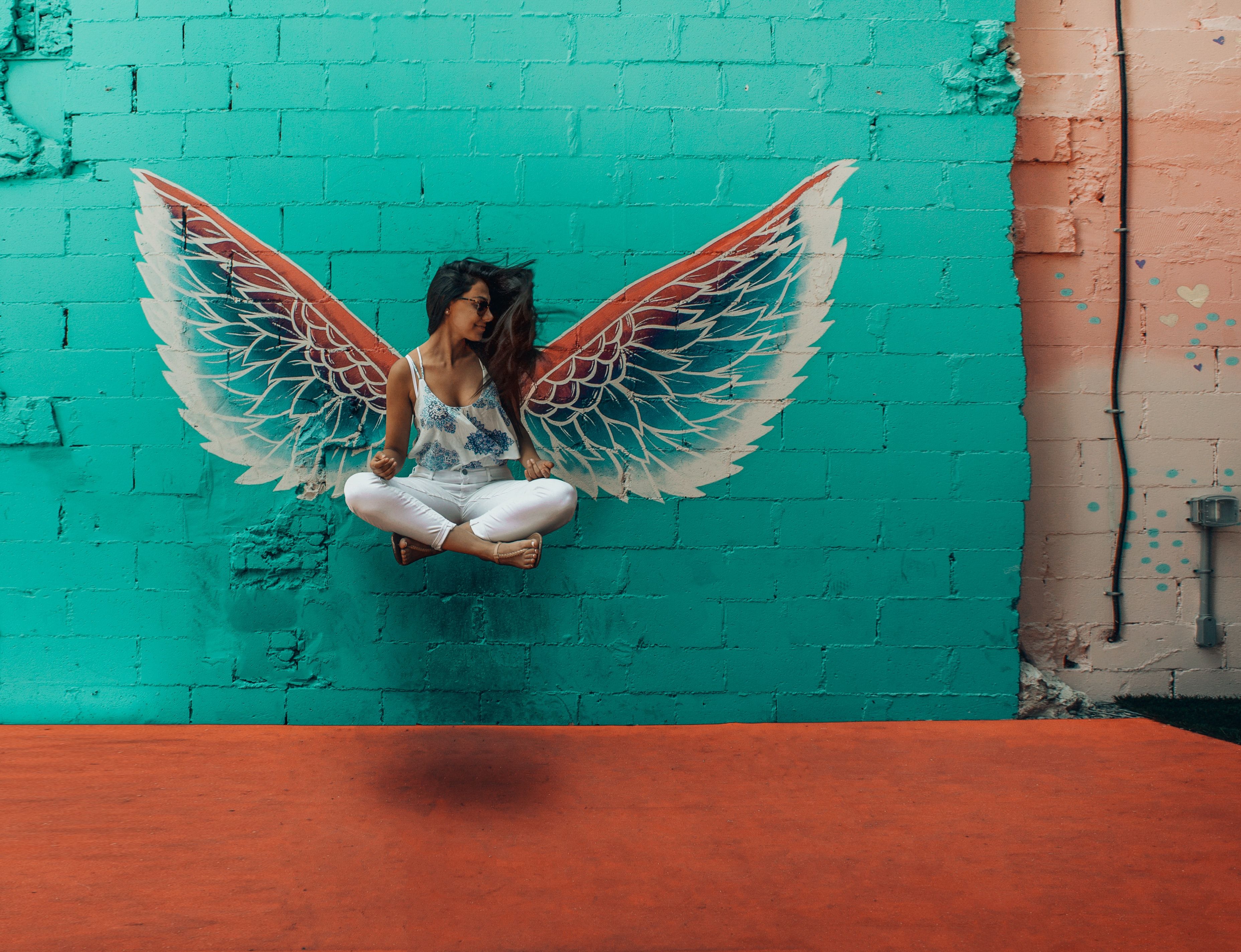 Woman with sunglasses sitting cross-legged against turquoise brick wall with painted angel wings