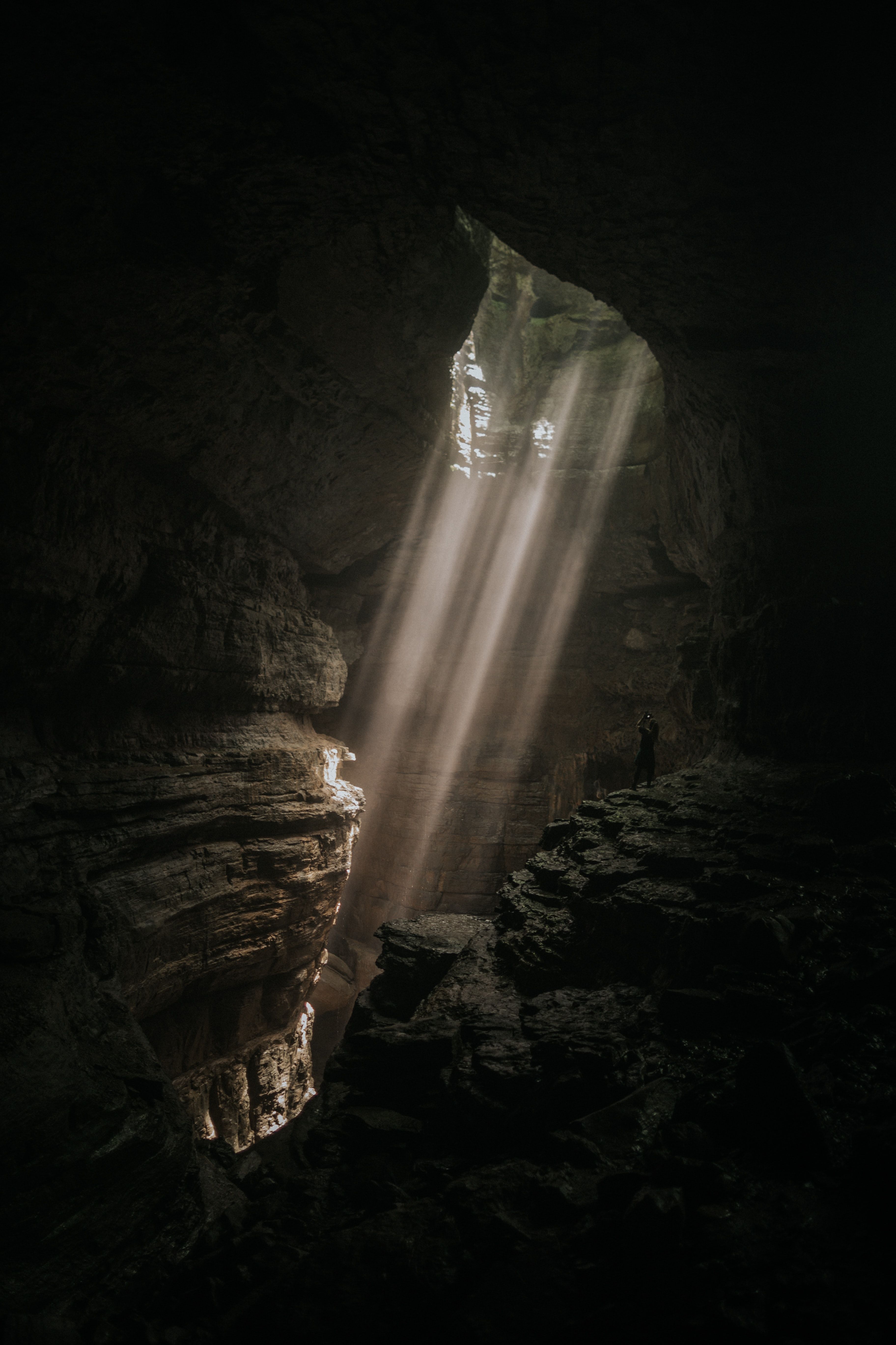 Sunlight streams through dark cave opening with rocky walls and a person standing silhouette