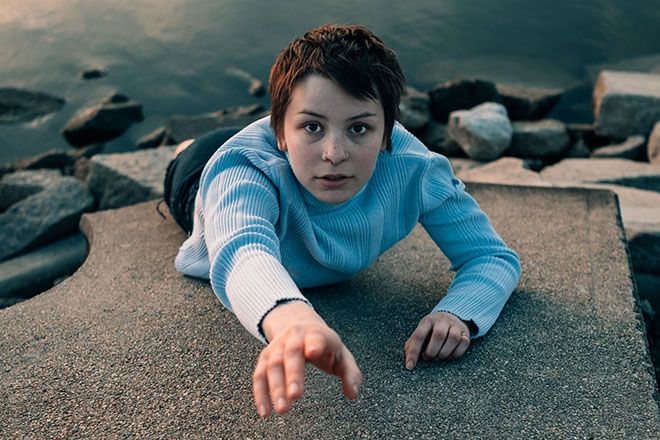 Young person in blue sweater lying on concrete by rocky waterfront, reaching forward