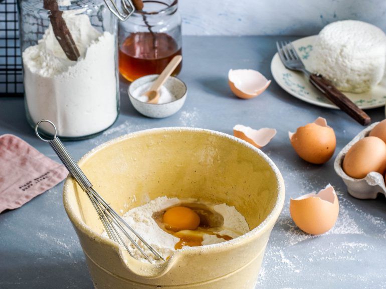 Baking ingredients and equipment arranged on surface with eggs, flour, whisk, and mixing bowl.