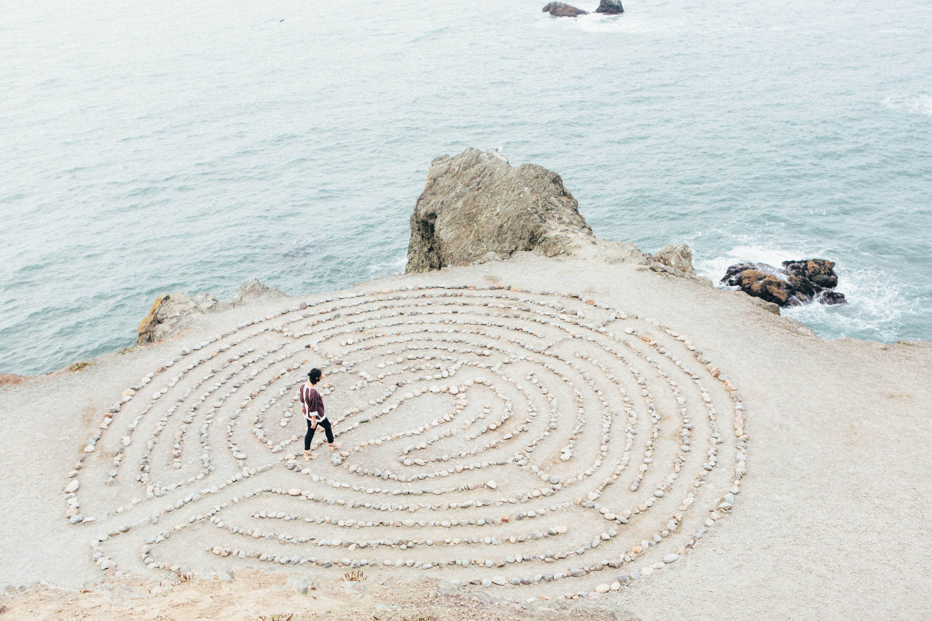 Person walking through spiral labyrinth made of stones on beach beside ocean rocks