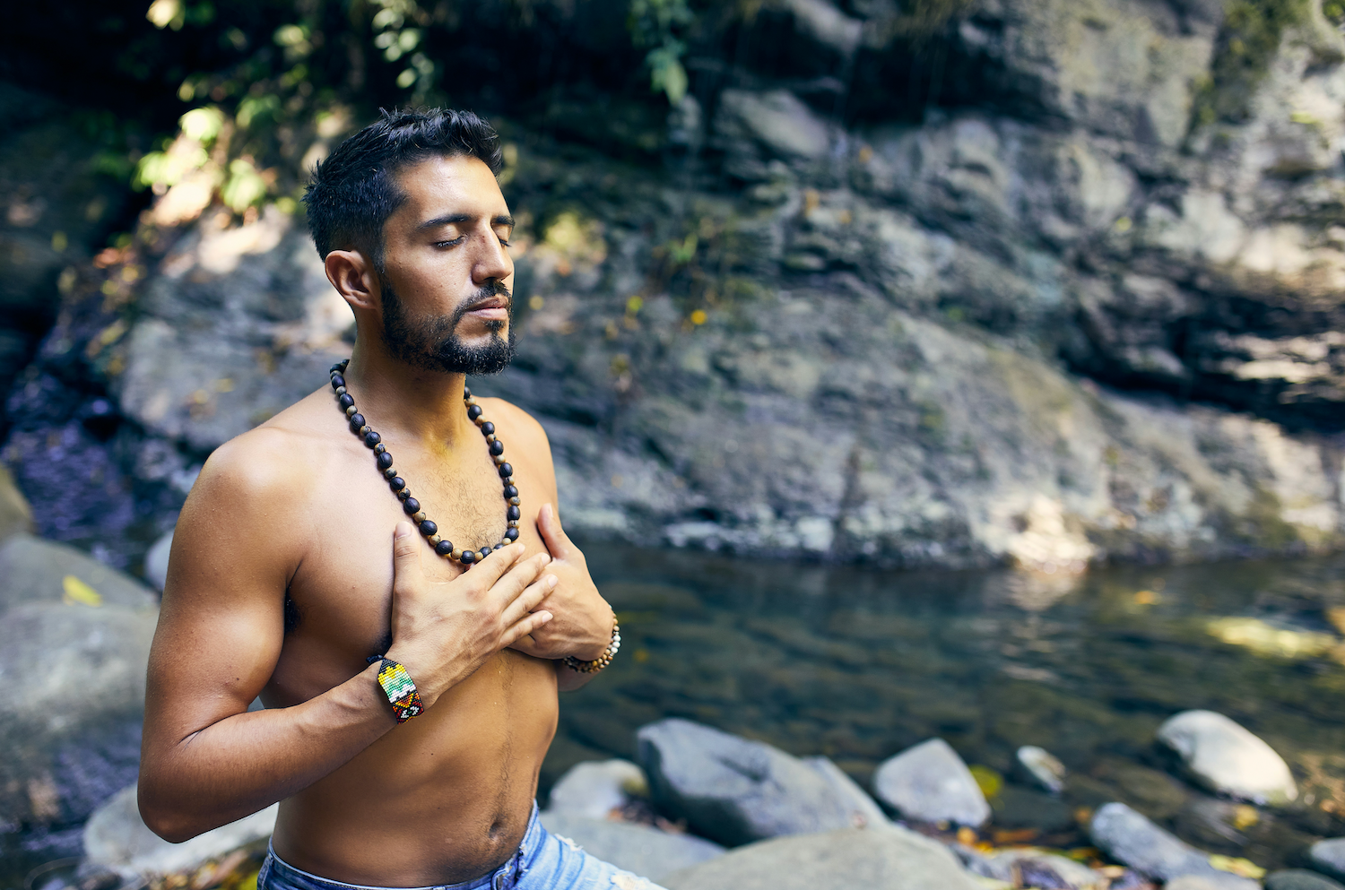 Shirtless man with beaded necklace standing thoughtfully by rocky stream.