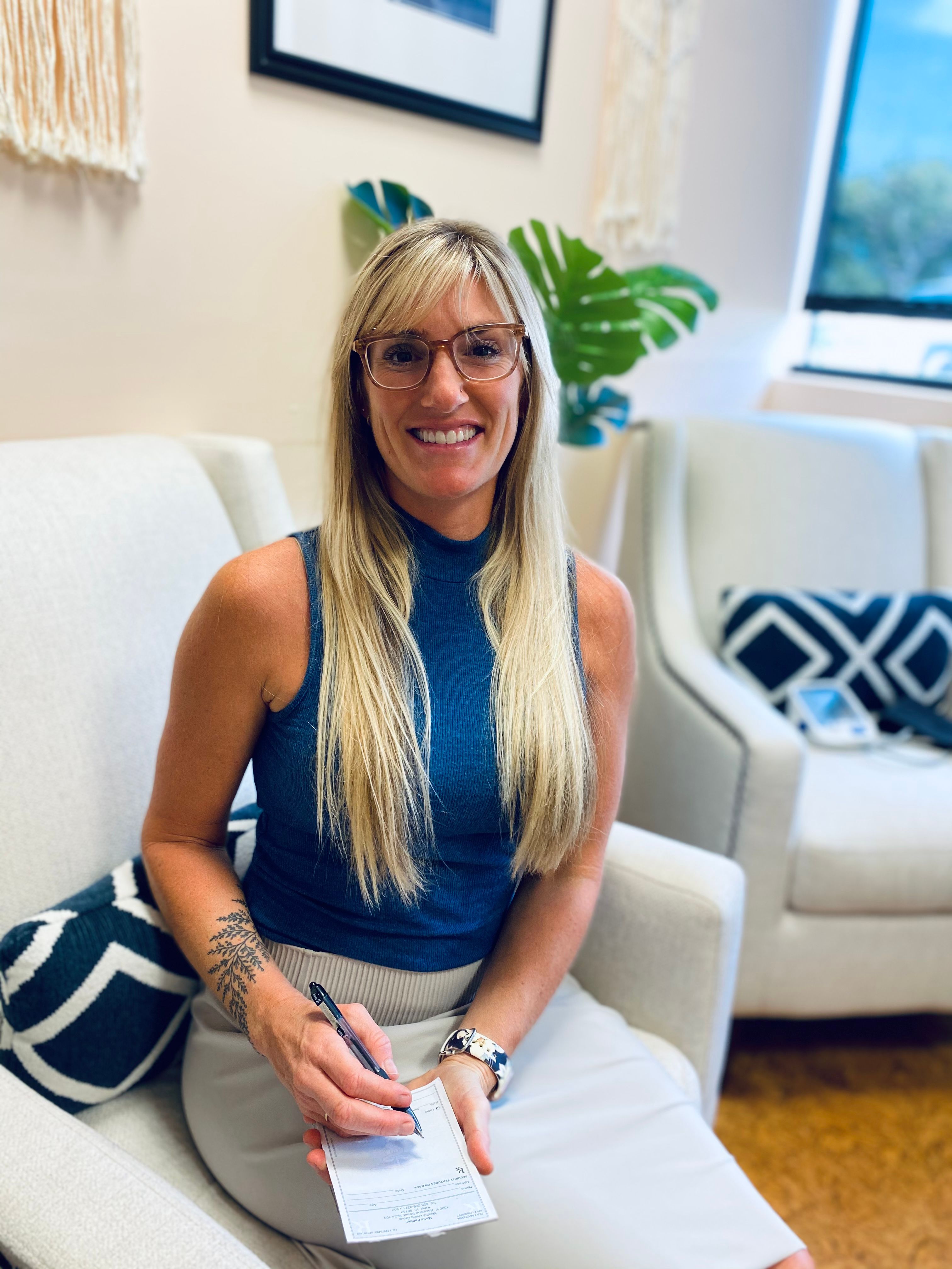 Smiling woman with blonde hair and glasses sitting on white couch holding clipboard