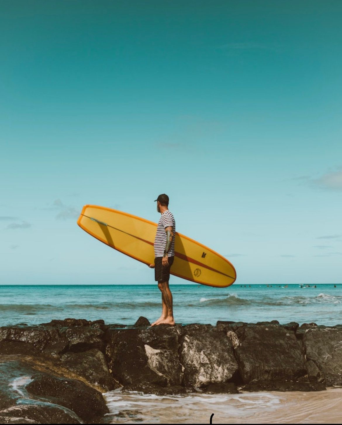 Man holding yellow surfboard standing on rocks at beach with turquoise ocean