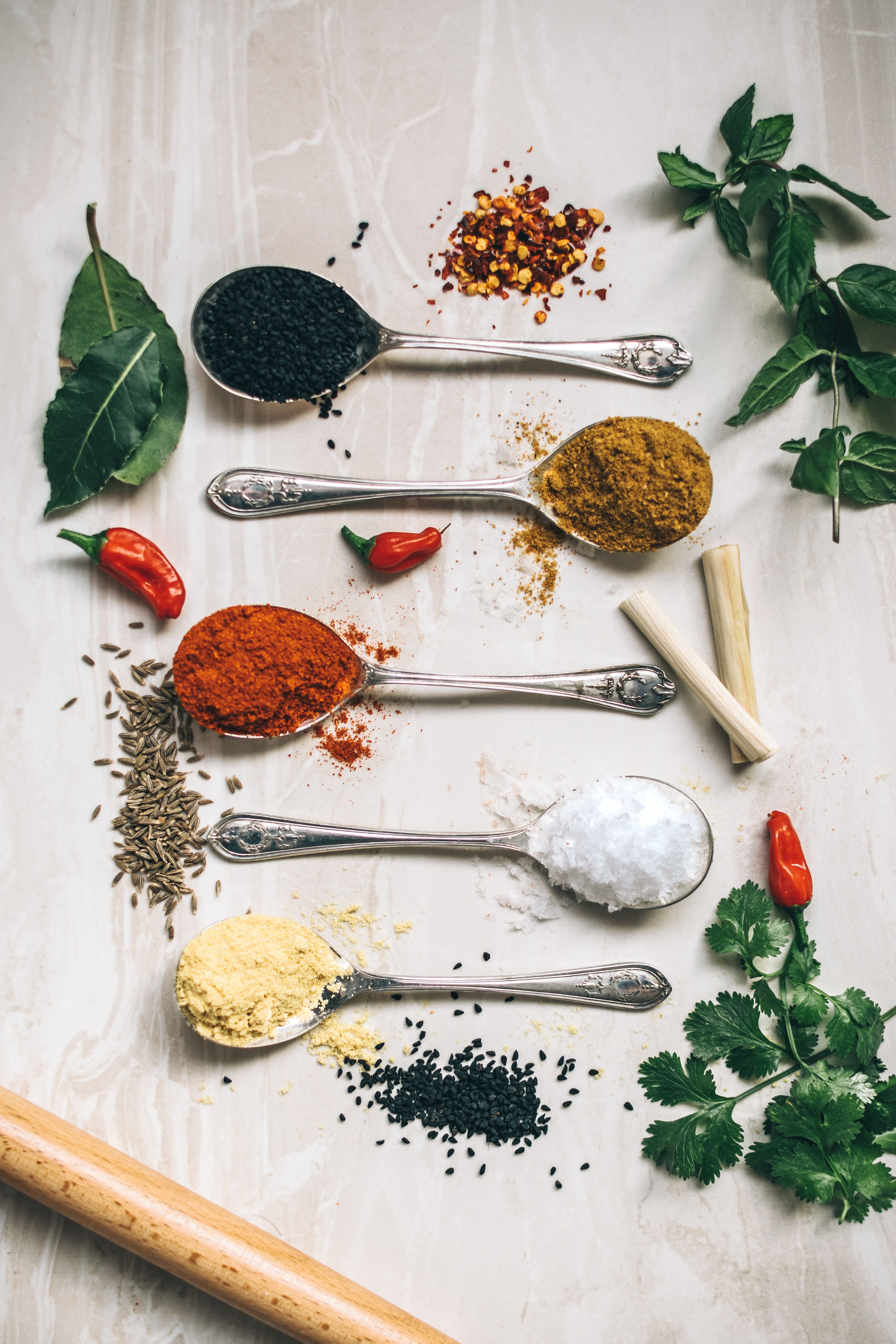 Spoons with various spices, herbs, and seasonings arranged on white textured surface
