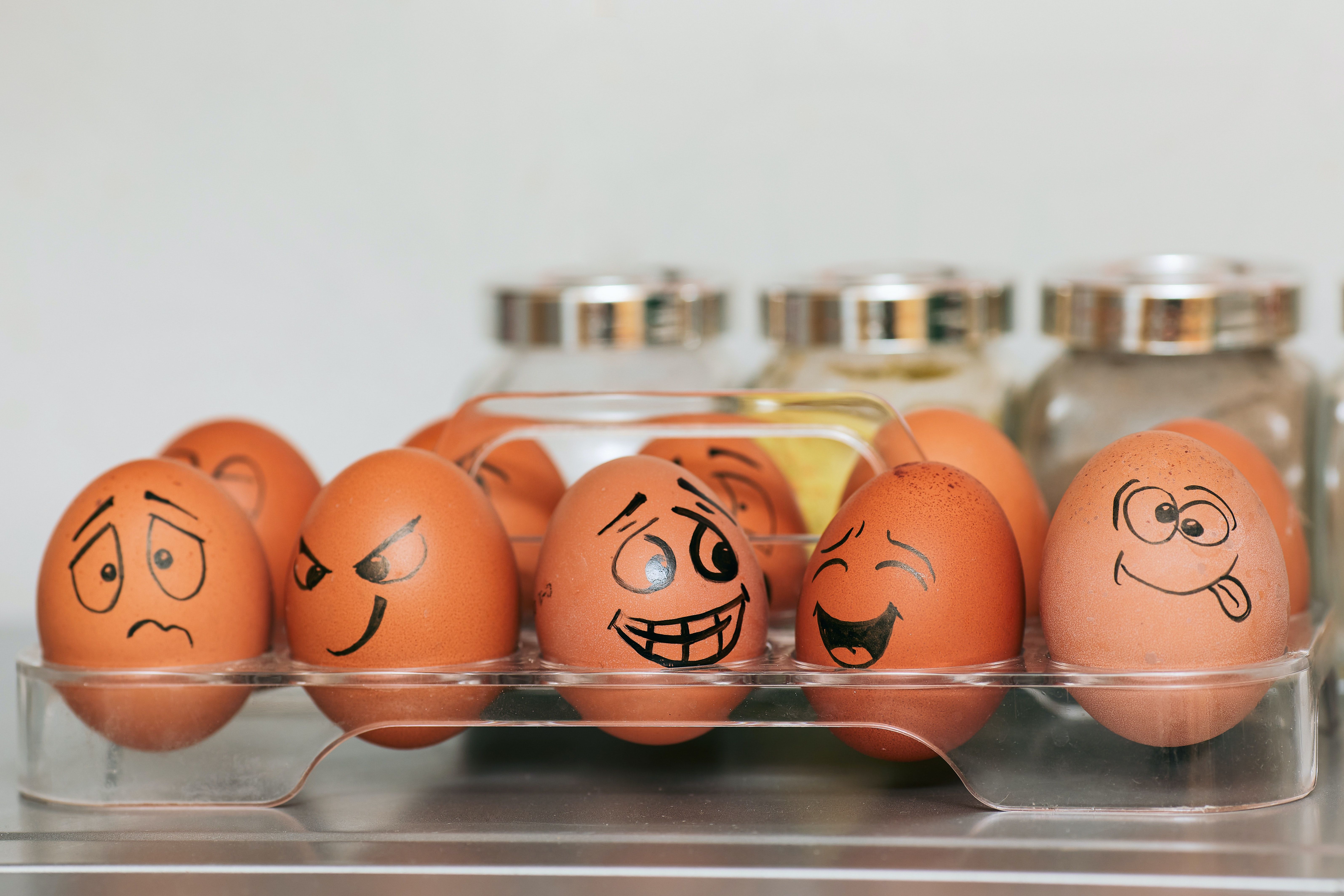 Five brown eggs with drawn faces showing different emotions in a clear plastic carton.