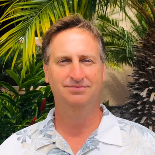 Middle-aged man in tropical shirt smiling outdoors with palm plants.