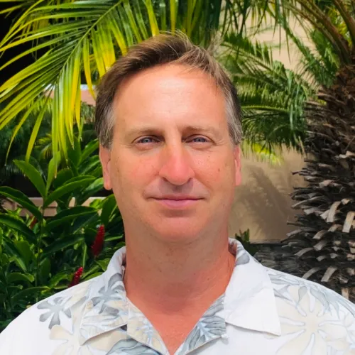 Middle-aged man in tropical shirt smiling outdoors with palm plants.