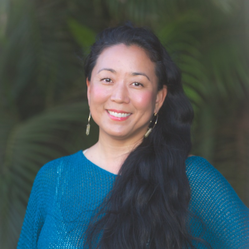 Woman with long black hair wearing blue textured top, smiling at camera with natural background