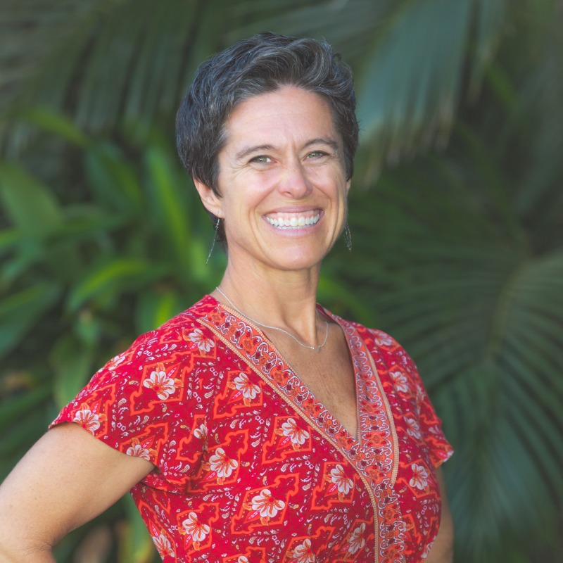 Woman with dark hair smiling, wearing red floral patterned shirt against green foliage background.