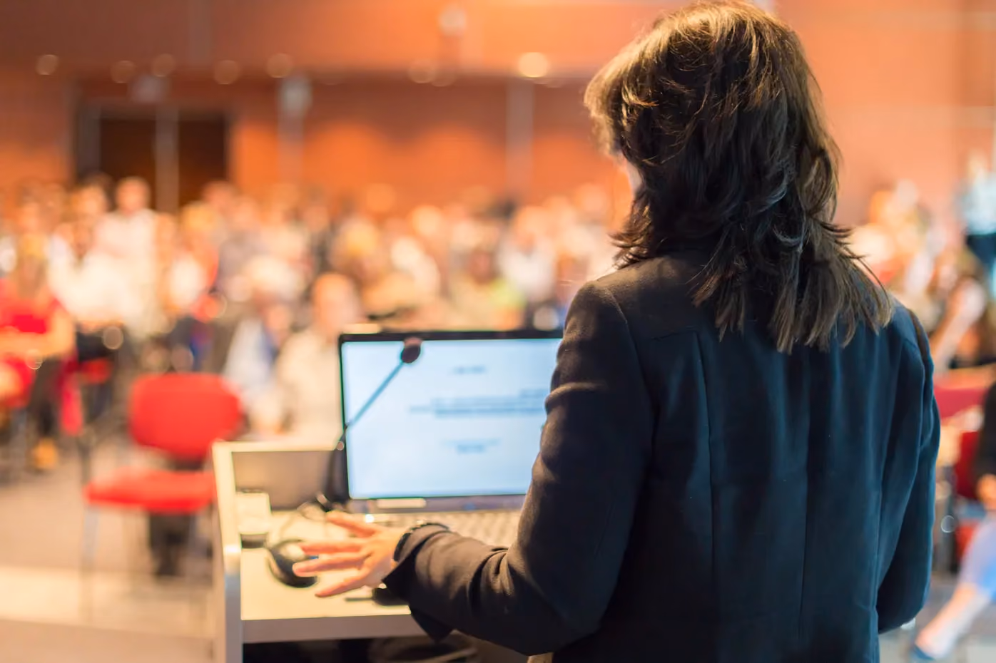 Woman speaker presenting to large audience at conference with laptop and microphone