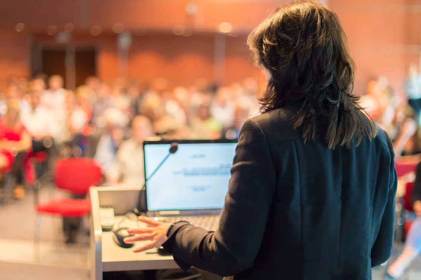 Woman speaker presenting to large audience at conference with laptop and microphone