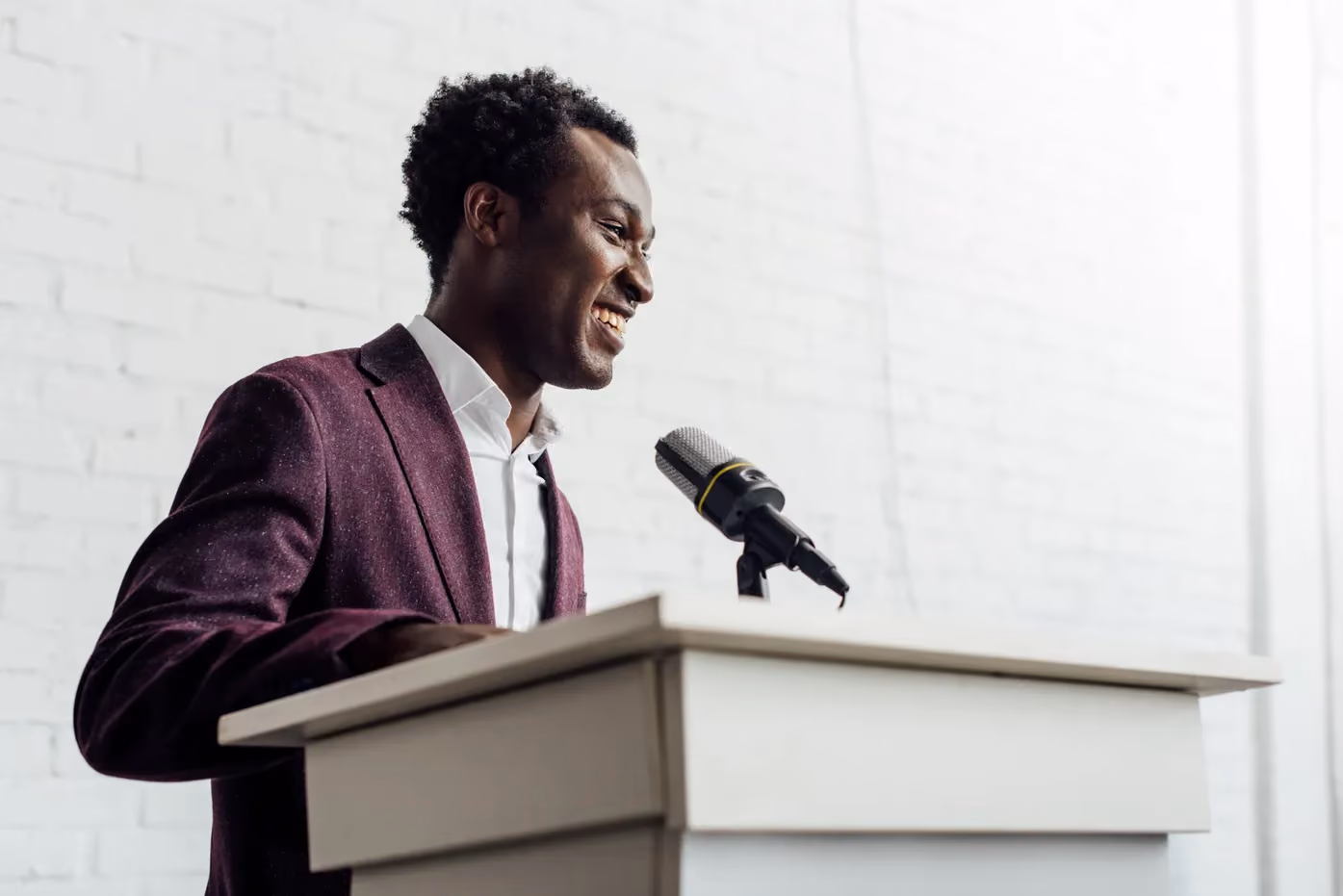 Man in burgundy blazer speaking at podium with microphone, smiling against white brick wall