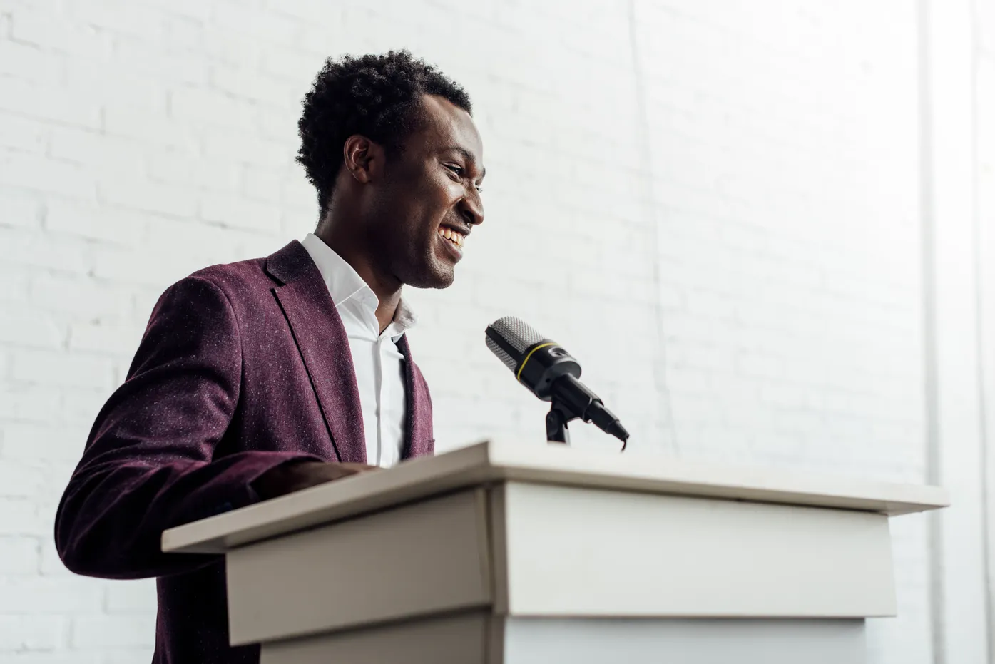 Man in burgundy blazer speaking at podium with microphone, smiling against white brick wall