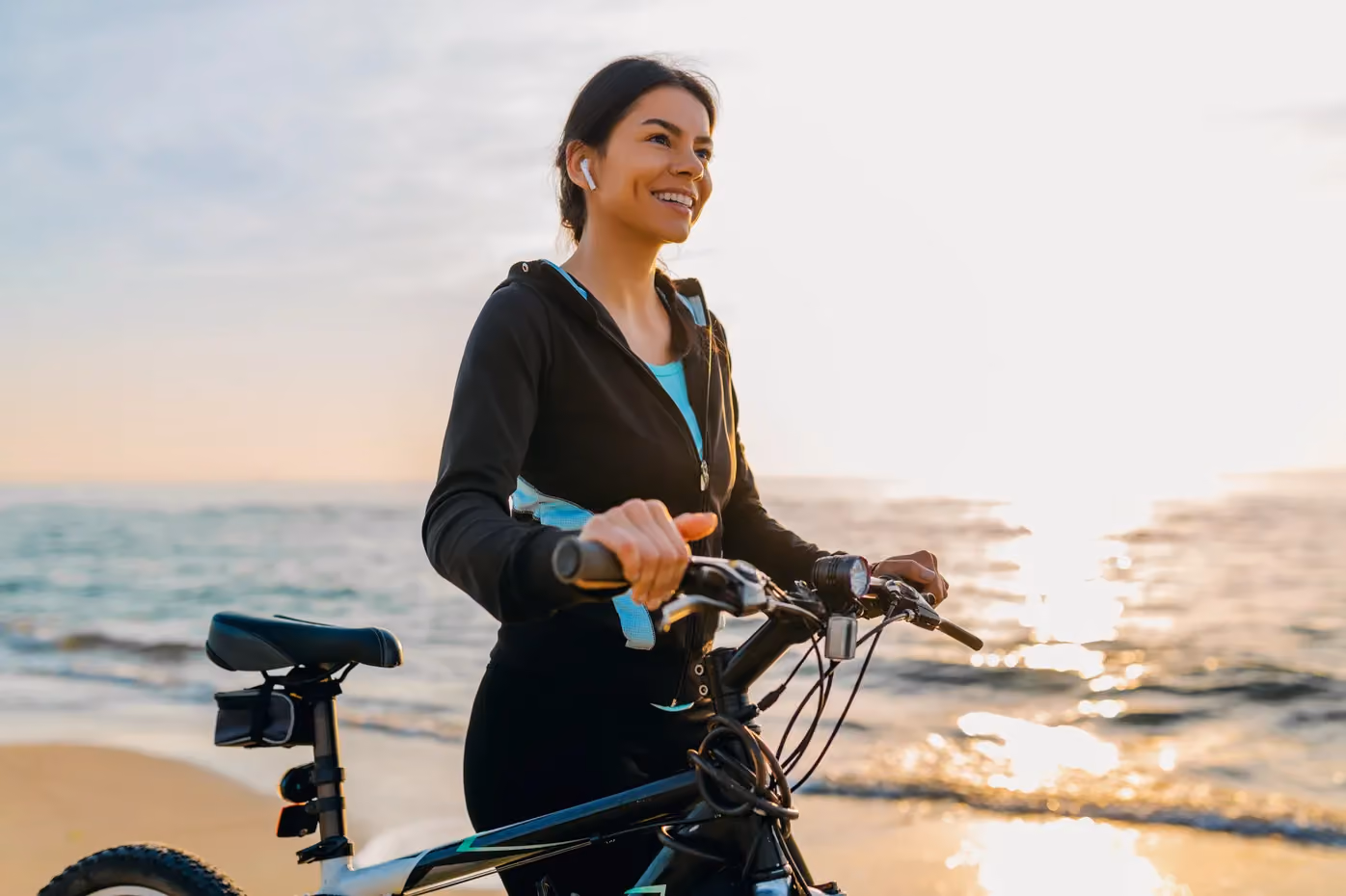 Woman riding bicycle on beach wearing earbuds and smiling at sunset