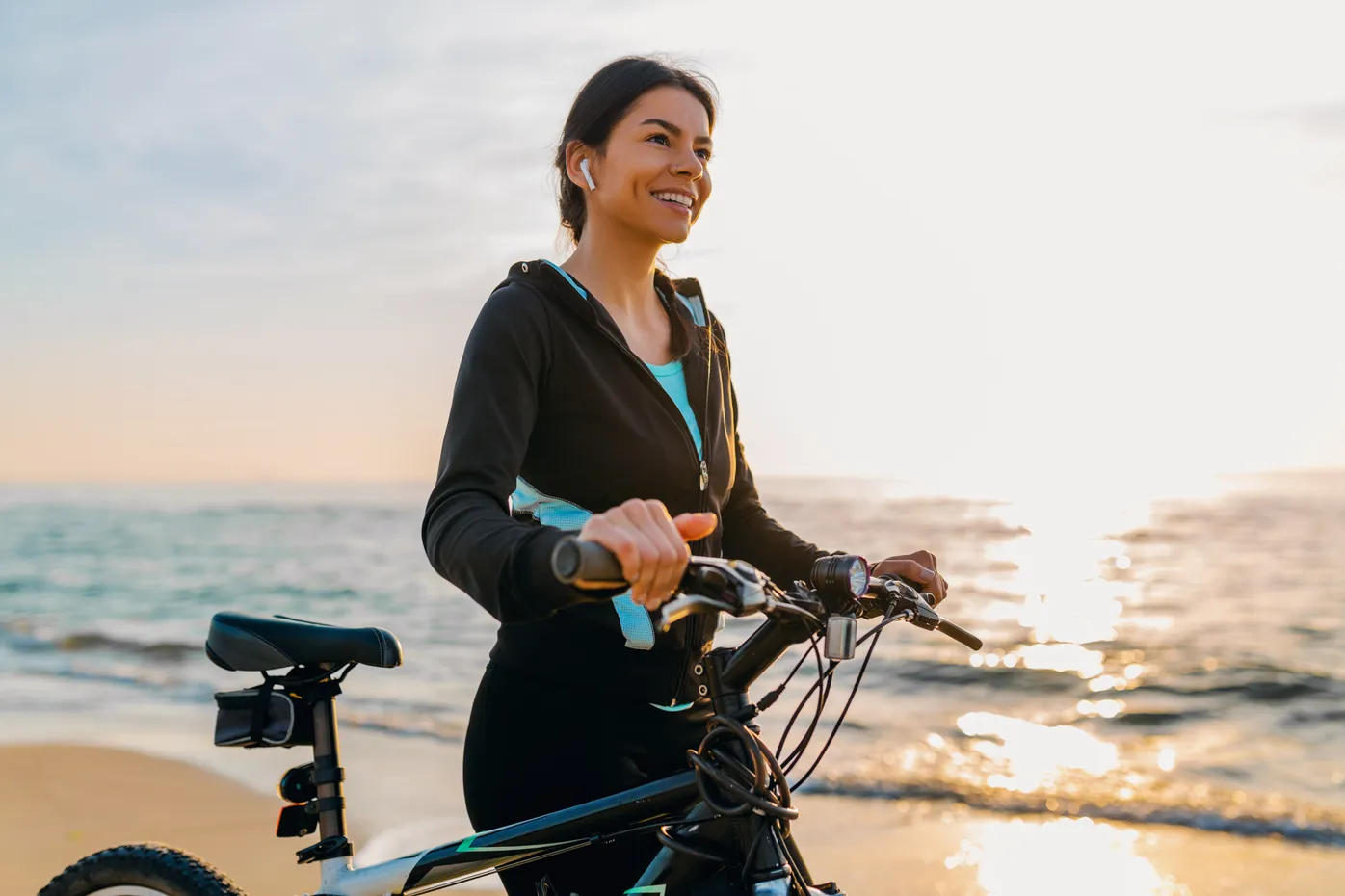 Woman riding bicycle on beach wearing earbuds and smiling at sunset