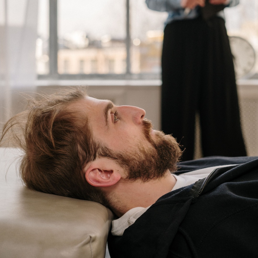 Man with brown beard reclining on couch, looking upward in modern living room.