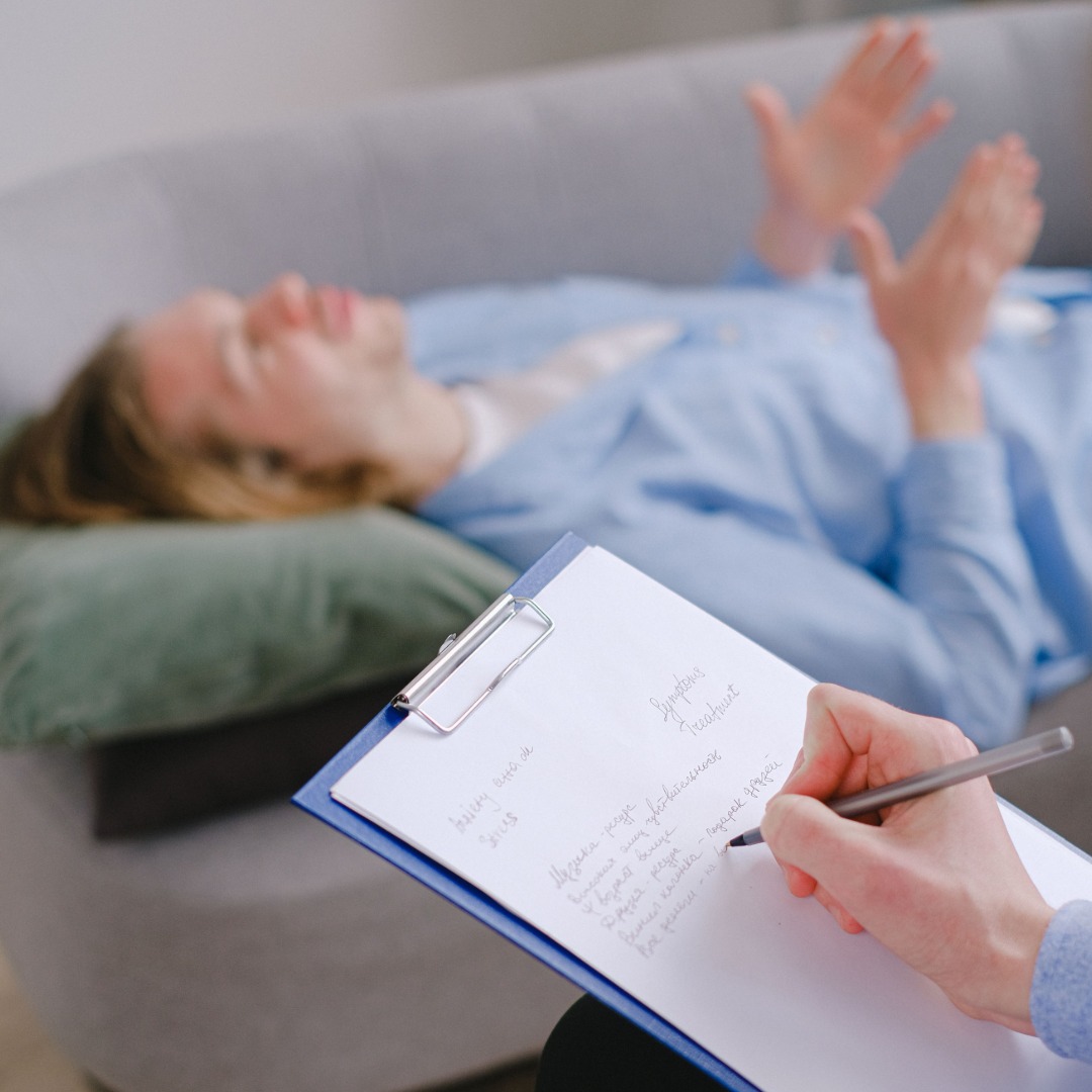 Therapist writing notes on clipboard while patient lies on couch during therapy session