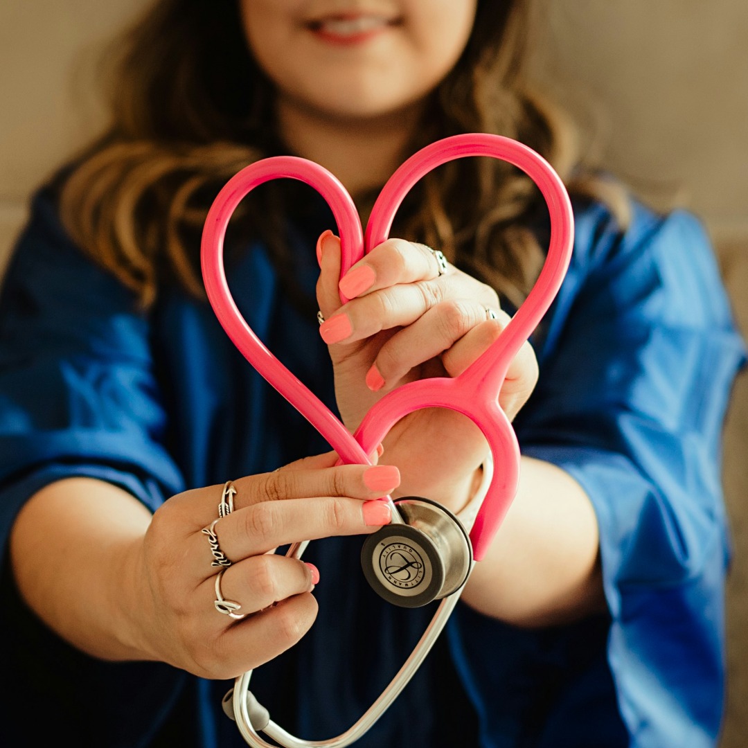 Woman in blue jacket holding pink heart-shaped stethoscope tube facing camera