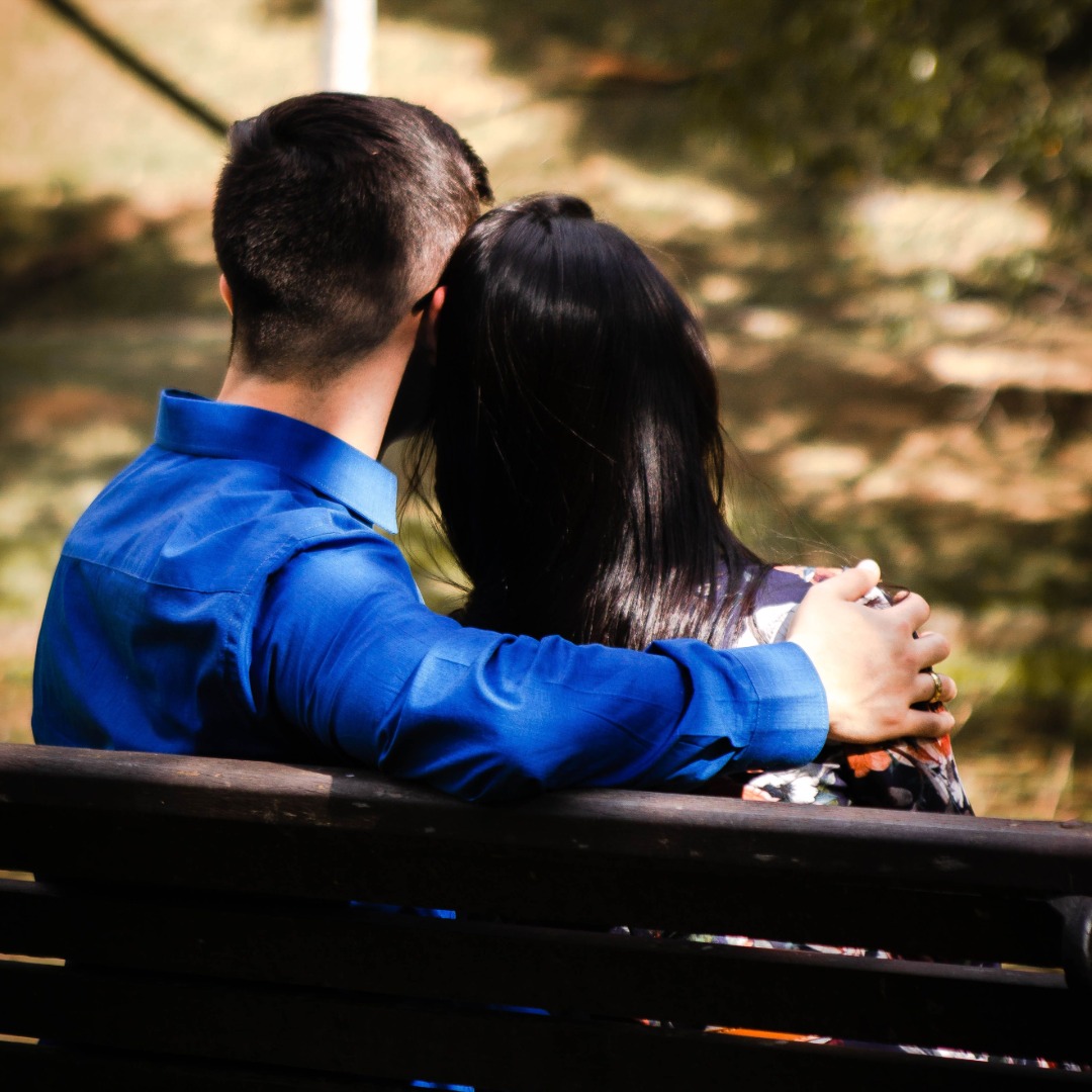 A couple sitting together on a bench outdoors, with the man wearing a blue shirt and woman with long dark hair