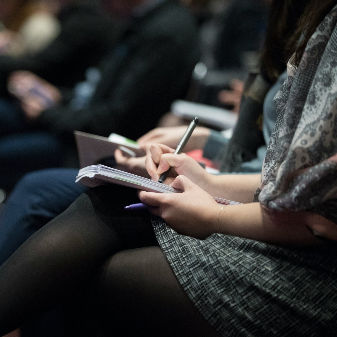 Person writing in notebook with pen during group meeting or event