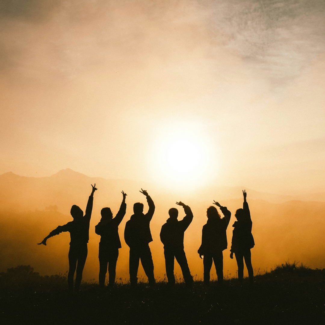 Silhouettes of six people with raised arms celebrating against golden sunset sky.