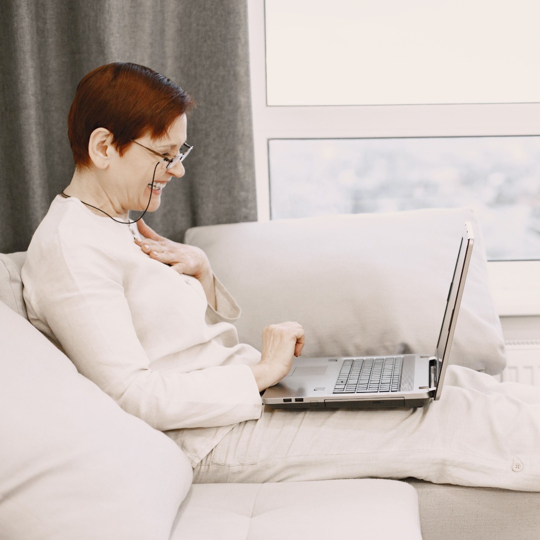 Middle-aged woman wearing glasses using laptop while sitting on beige couch near window