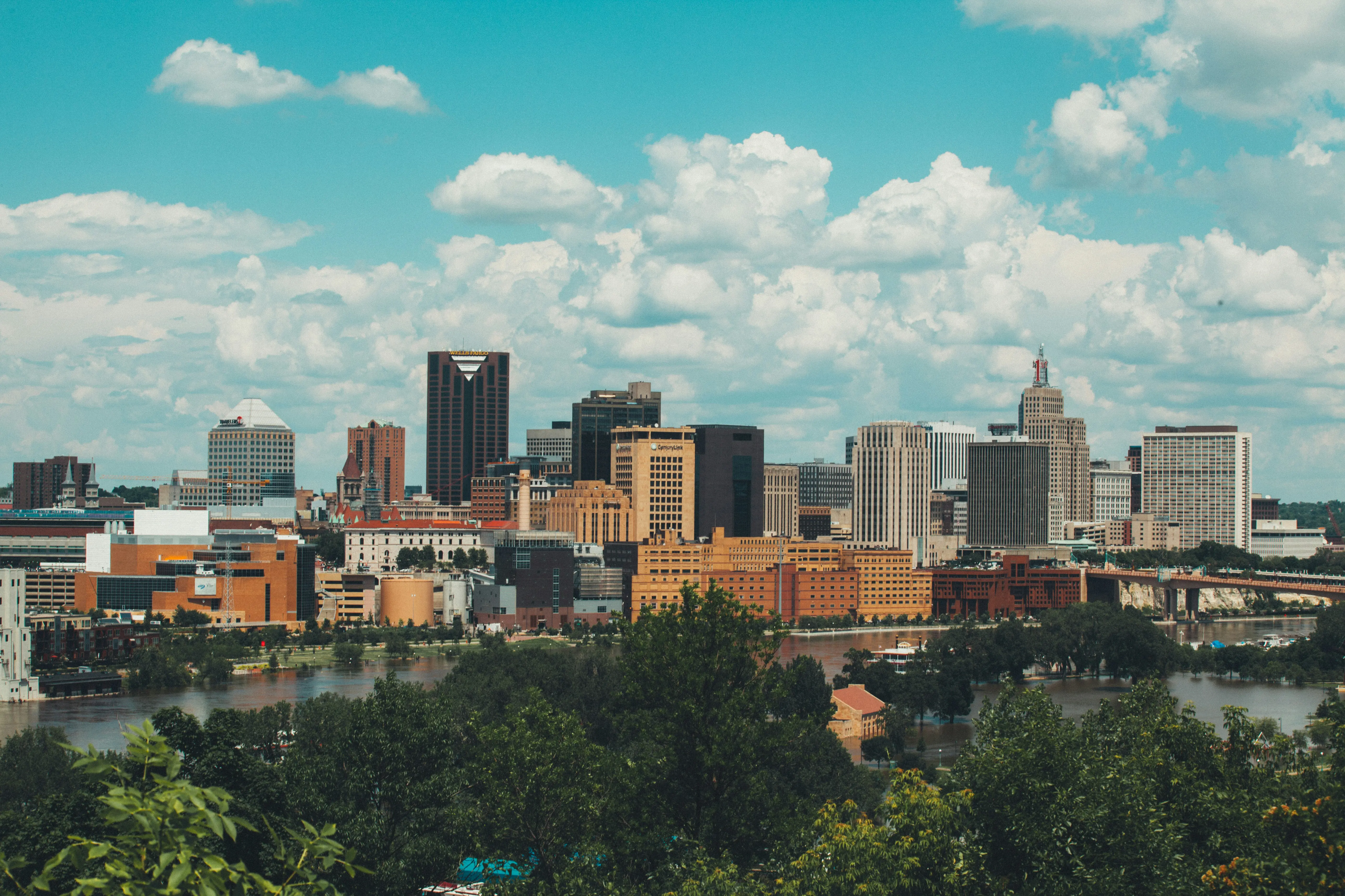 City skyline with diverse buildings under a blue sky with scattered clouds and trees in the foreground.