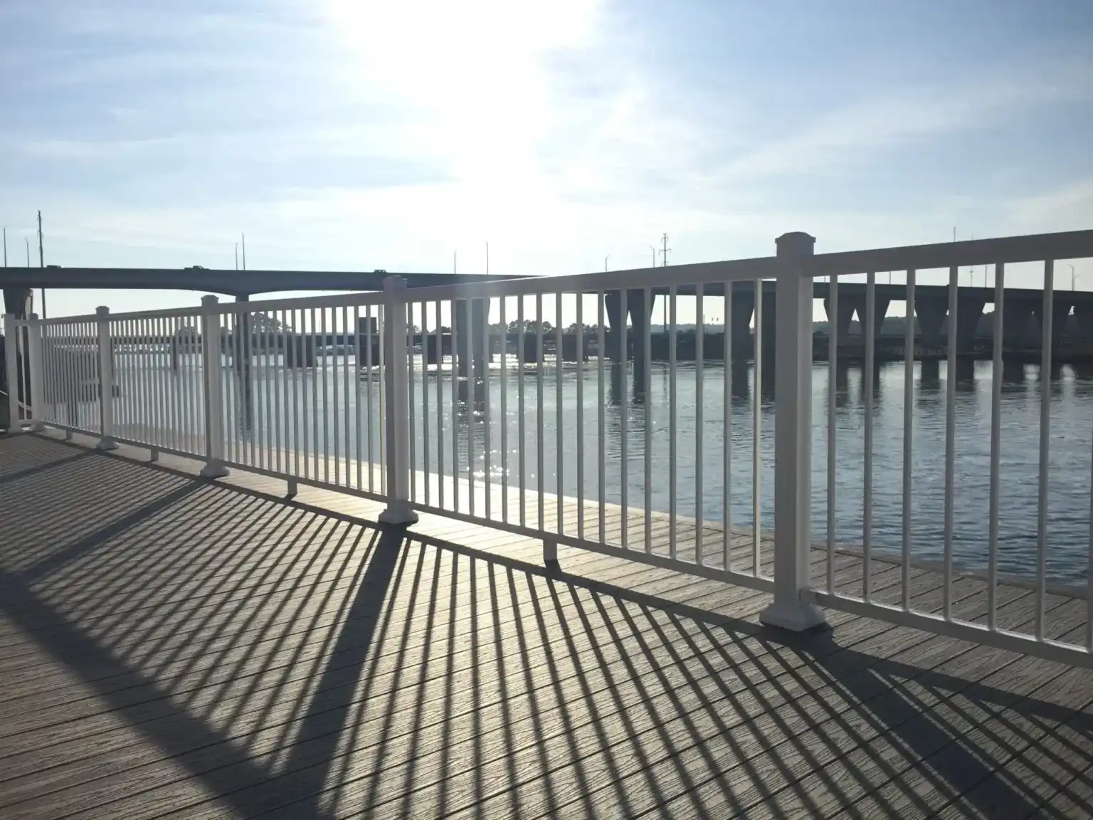 White metal fence casting shadows on wooden deck with river and bridge in bright sunlight.