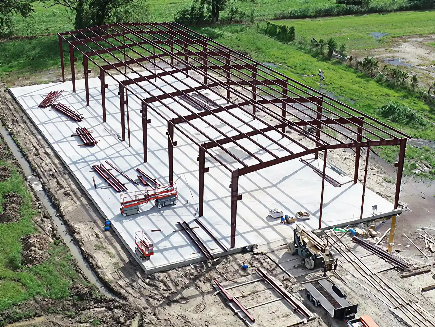 Steel frame structure under construction on a concrete slab surrounded by grass and dirt.