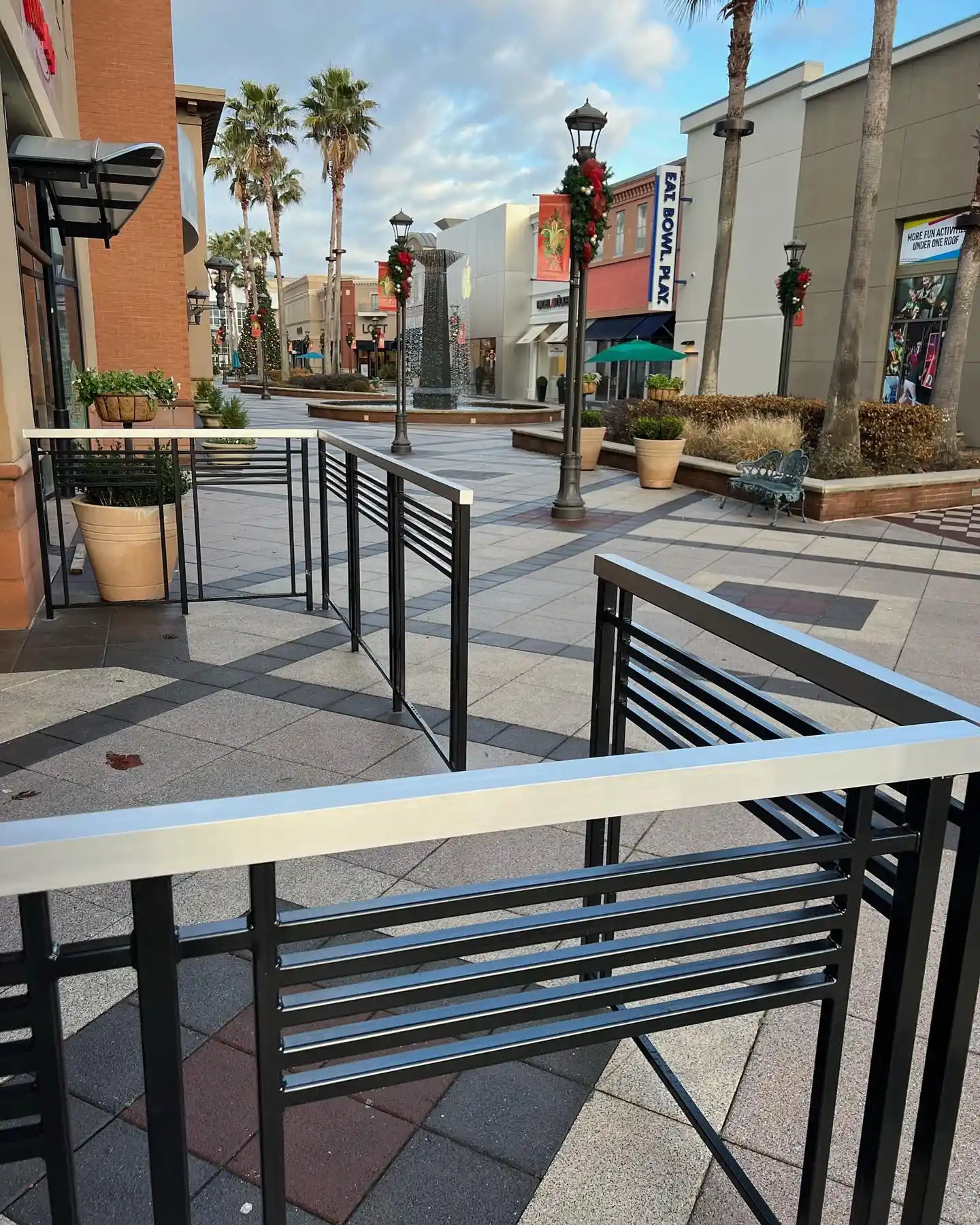Outdoor shopping area with decorative railings, palm trees, lampposts with wreaths, and a fountain in the distance.