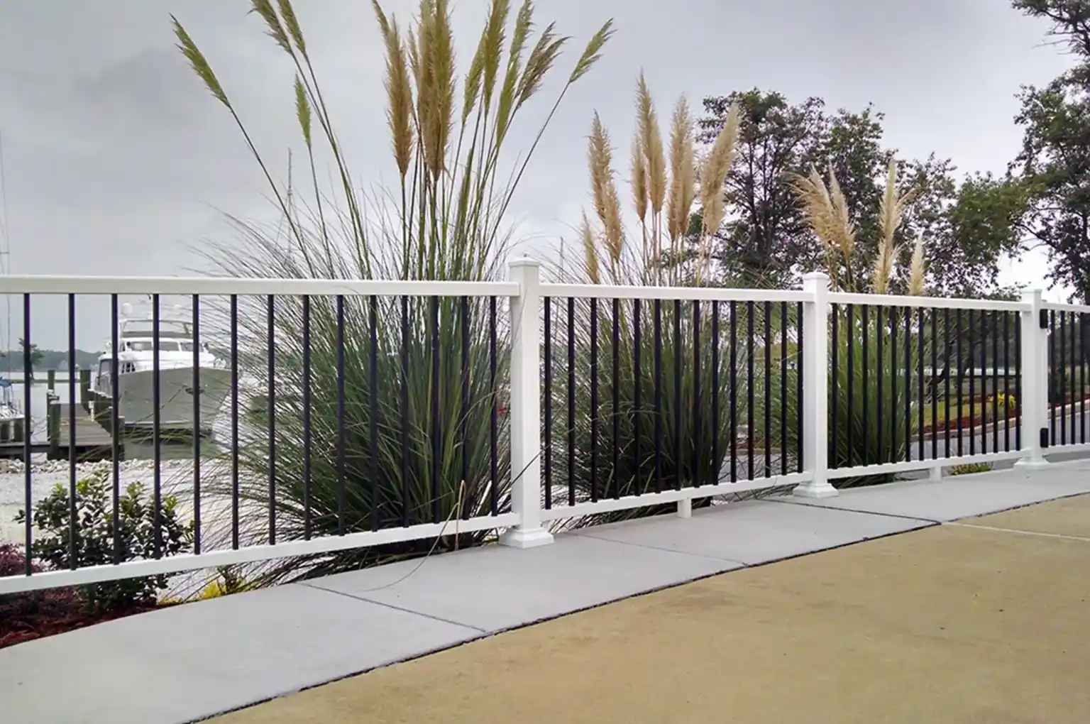 White and black metal fence along a sidewalk with tall ornamental grasses and a docked yacht in background.