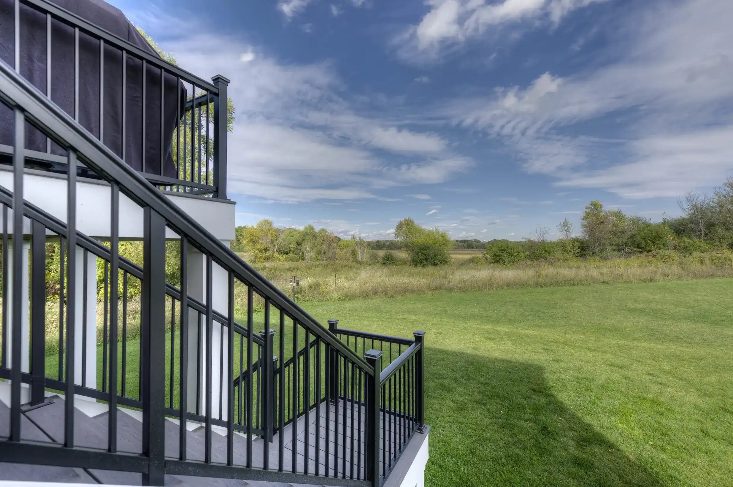 Outdoor staircase with black railing overlooking a grassy field and trees under a partly cloudy sky.