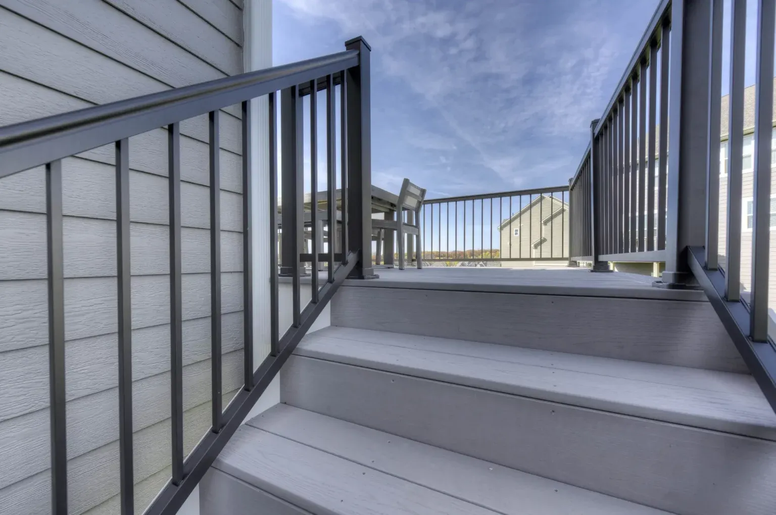 Gray outdoor stairs with black metal railing leading to a balcony with a table and two chairs.