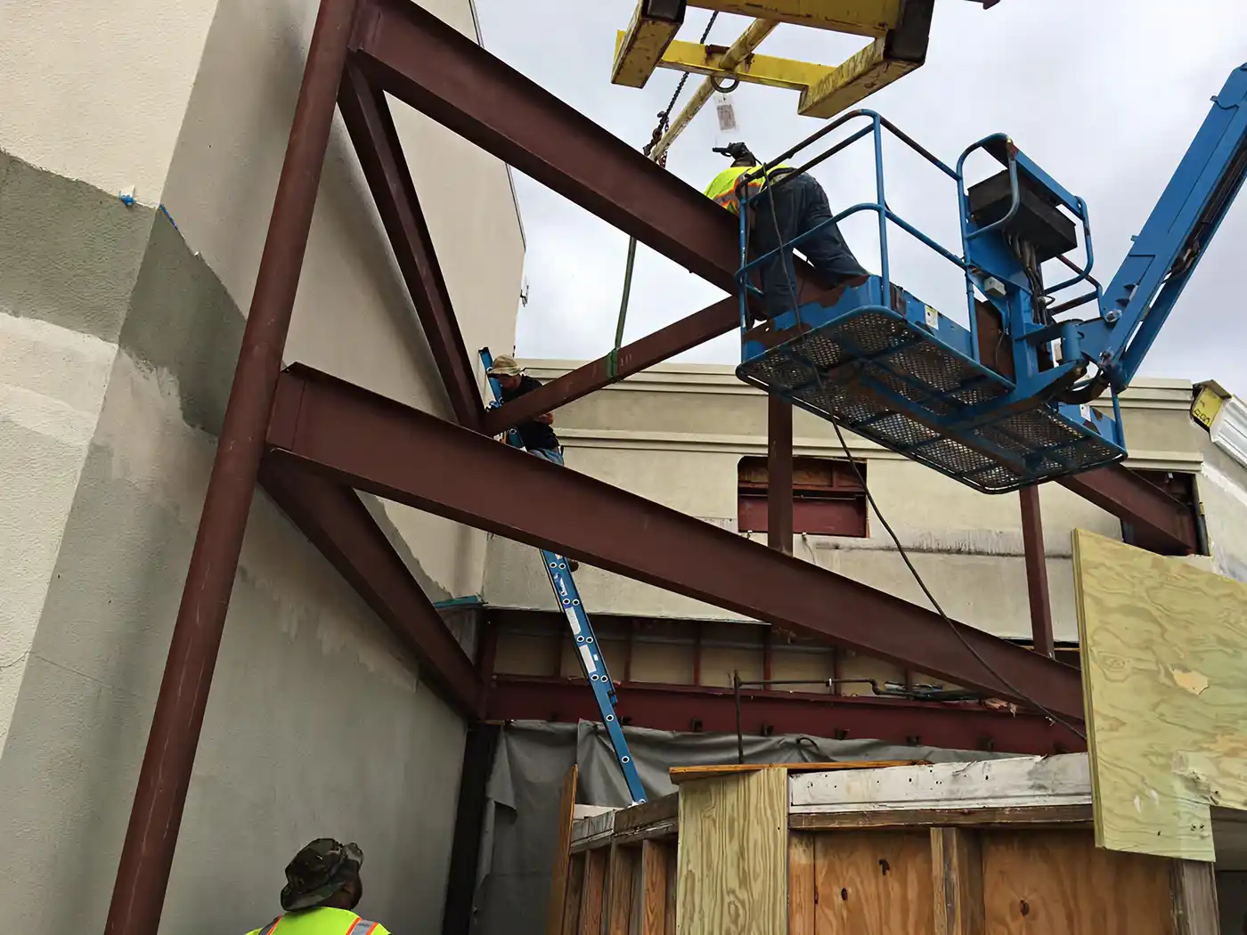 Construction workers on ladders and lift installing large steel beams on a building exterior.