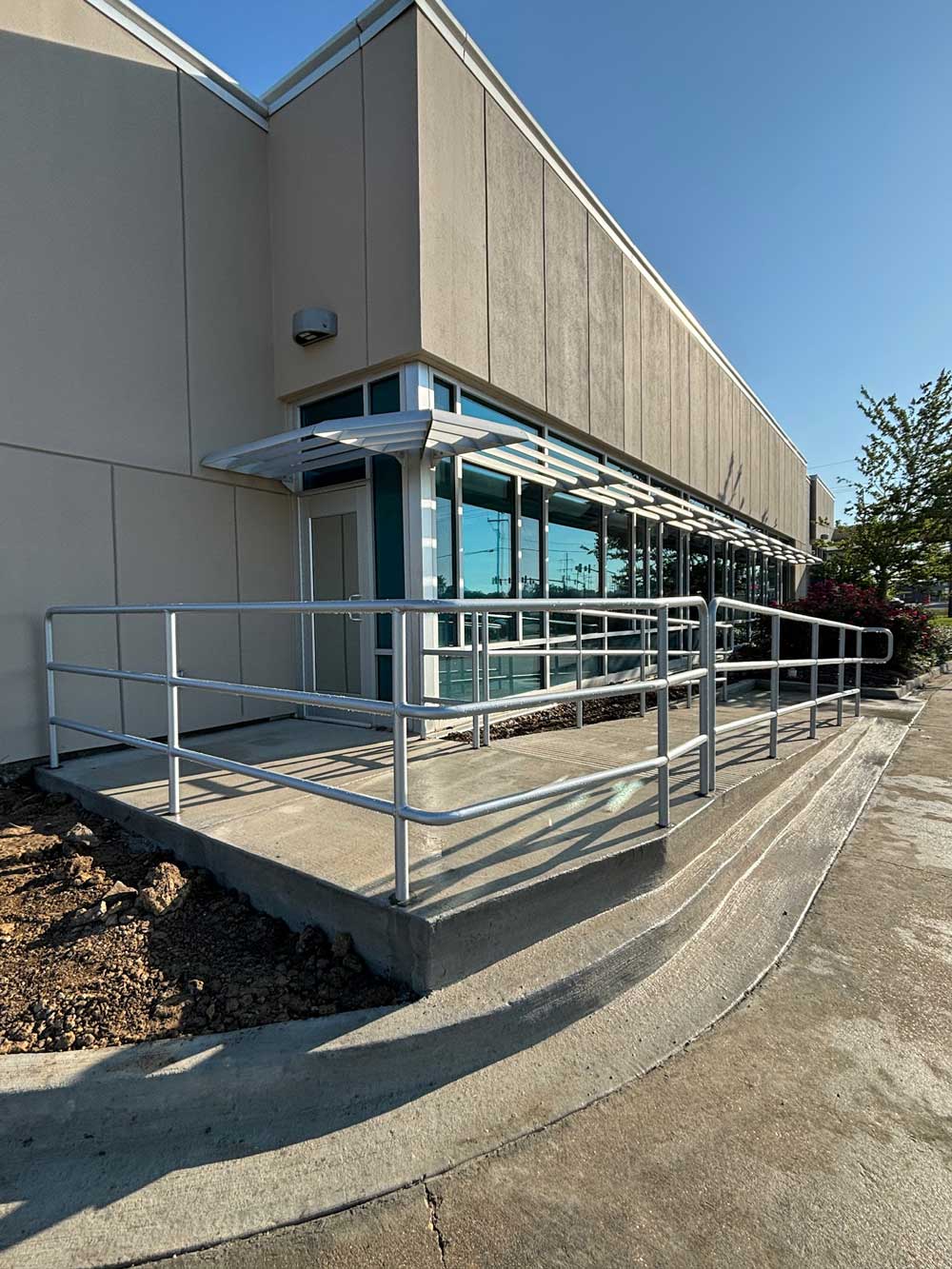 Concrete wheelchair ramp with silver metal railings leading to a glass doorway on a beige building.