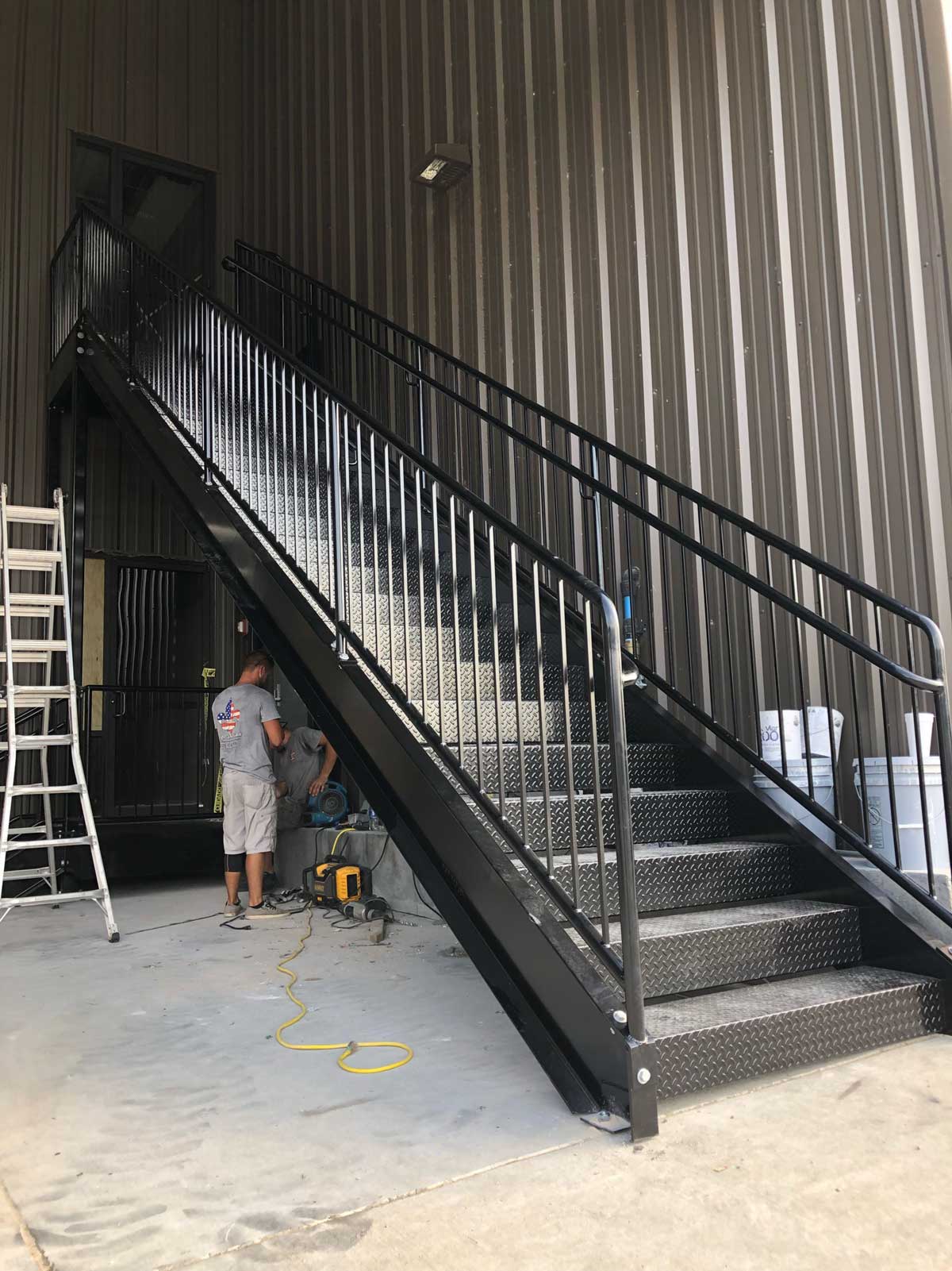 Two workers near a black metal staircase inside an industrial building with corrugated walls.